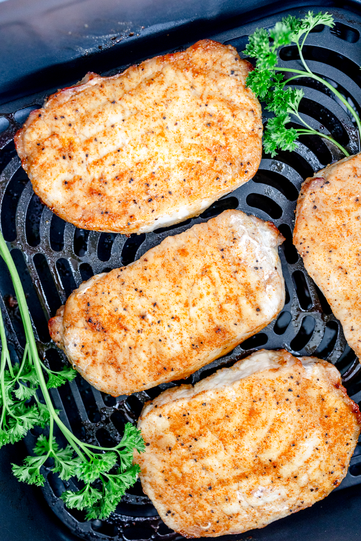 Close-up overhead view of four fully cooked seasoned pork chops, golden-brown in color, placed in an air fryer basket garnished with fresh parsley.