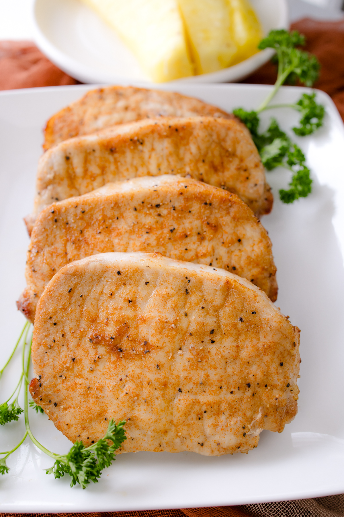 Angled overhead view of four cooked, seasoned pork chops served whole on a square white plate garnished with fresh parsley.
