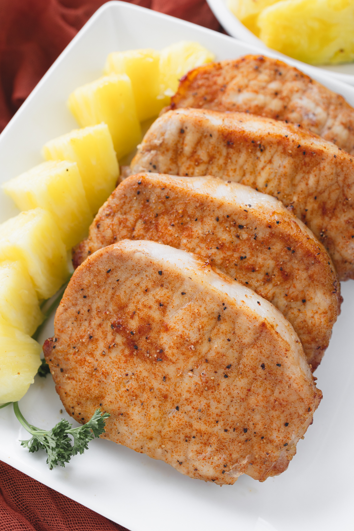 Close-up angled overhead view of four cooked seasoned pork chops served whole on a square white plate with a fresh parsley garnish.