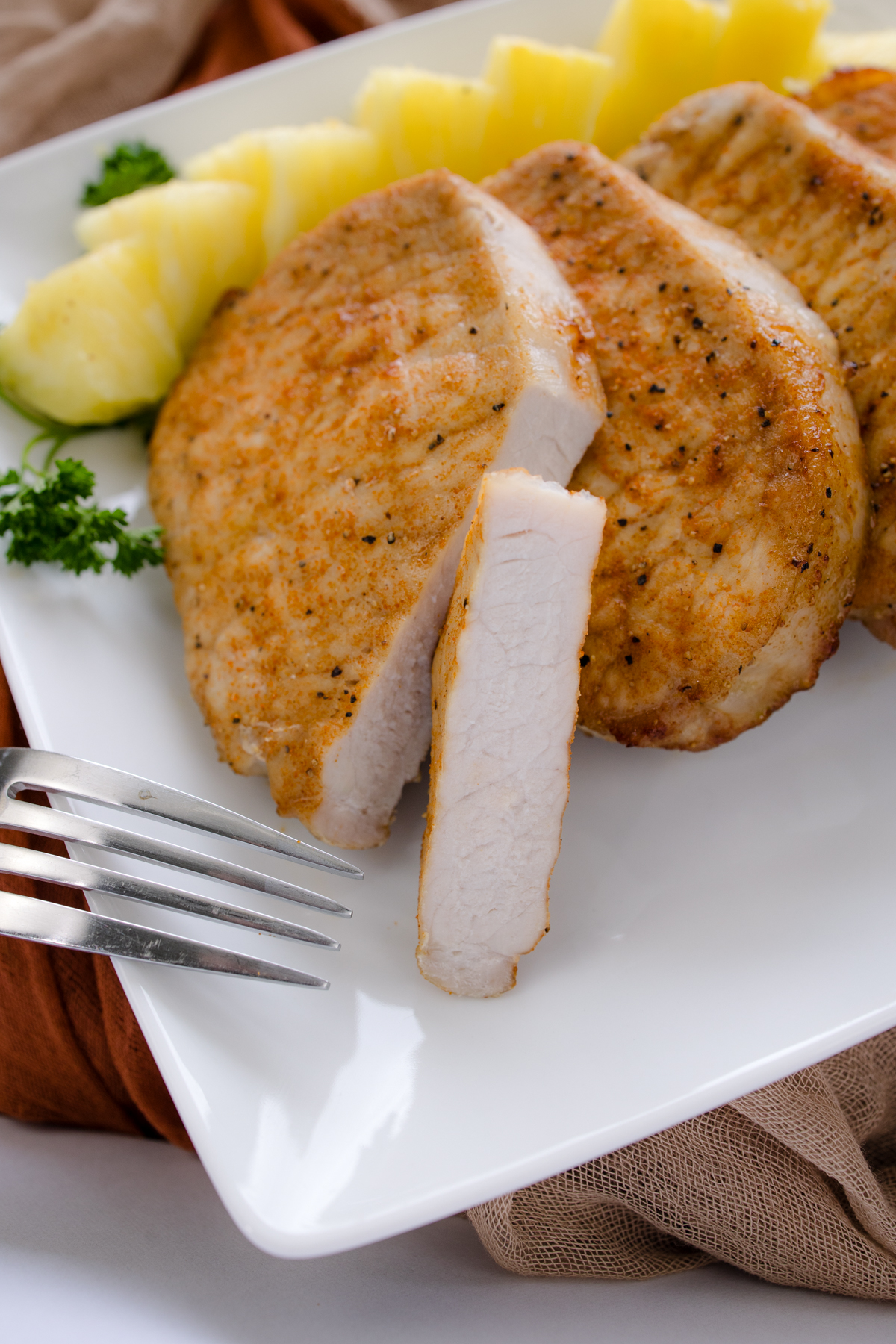 Close-up angled overhead view of a pork chop, sliced to clearly show juicy texture and crispy edges, on a white plate next to a fork and stacked beside other pork chops.