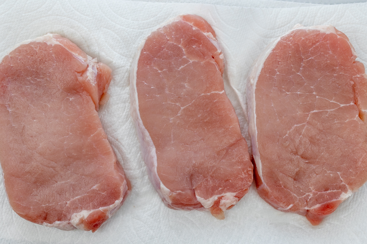 Overhead view of three raw, unseasoned pork chops arranged neatly on white paper towel.