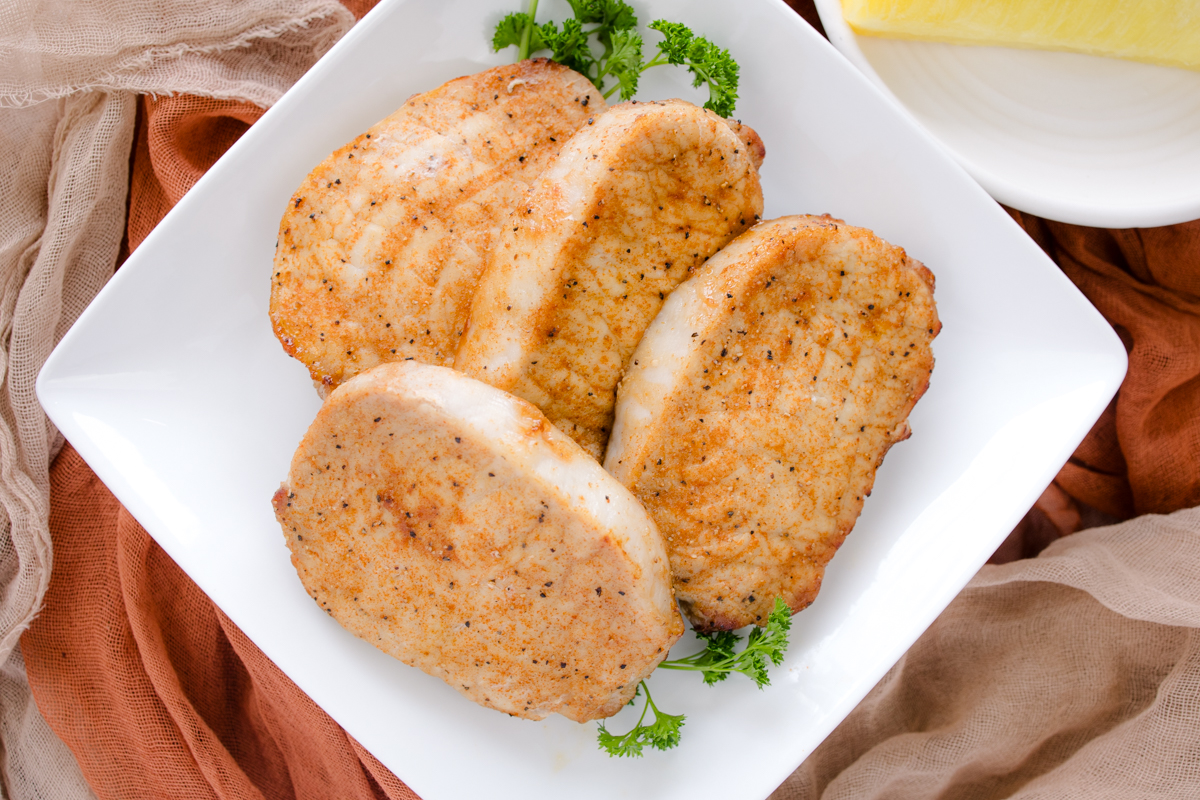 Close-up angled overhead view of four cooked seasoned pork chops served whole on a square white plate with a fresh parsley garnish.