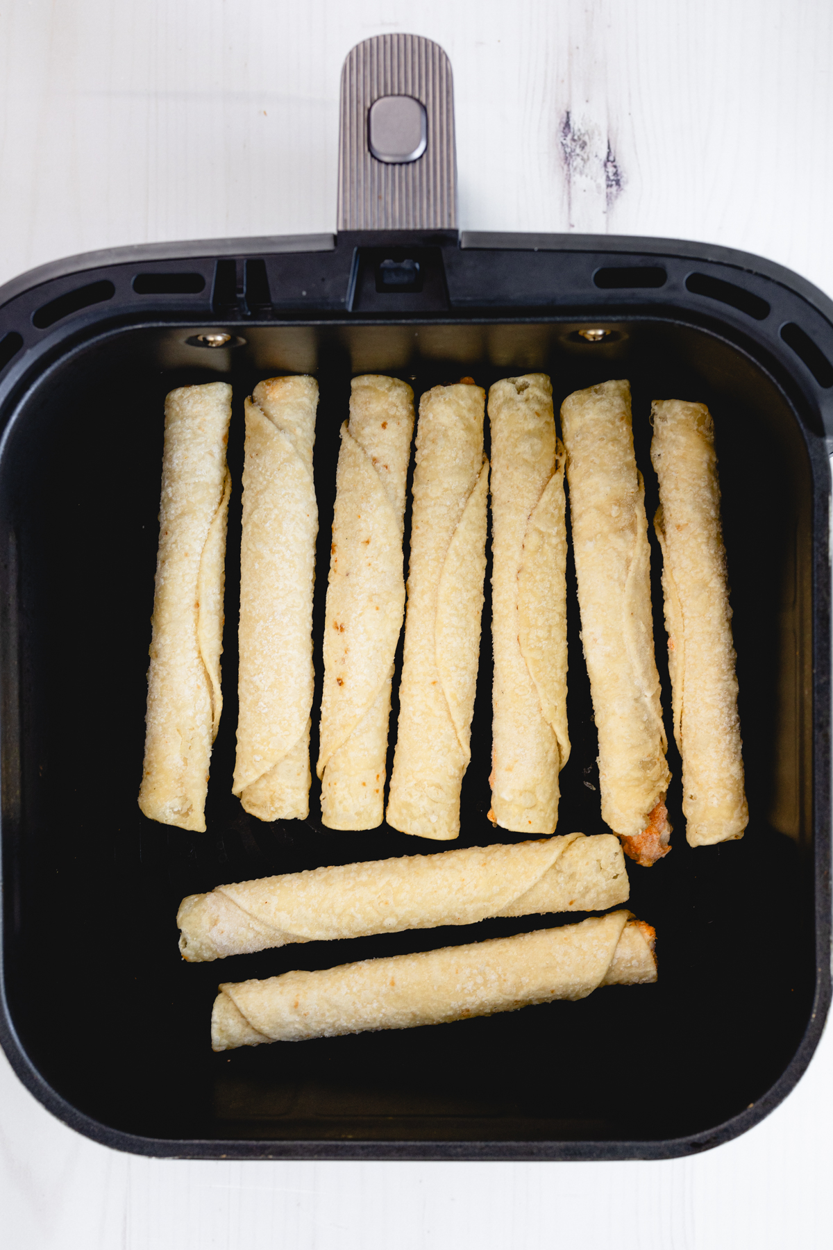 Overhead view of nine uncooked chicken taquitos arranged in an air fryer basket.