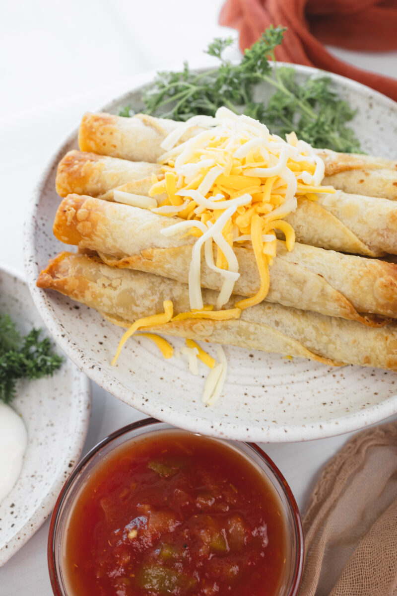 Overhead view of crispy chicken taquitos stacked on a plate with shredded cheese, fresh parsley, and a small dish of salsa.