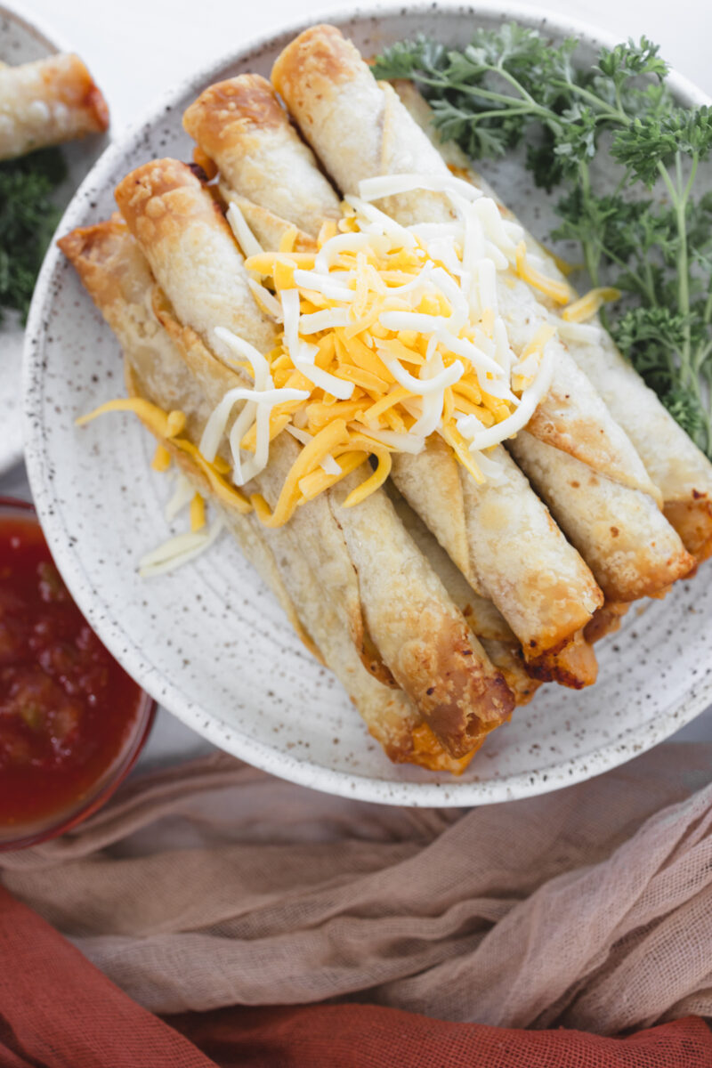 Close-up overhead view of a plate stacked with chicken taquitos topped with shredded cheese and parsley.