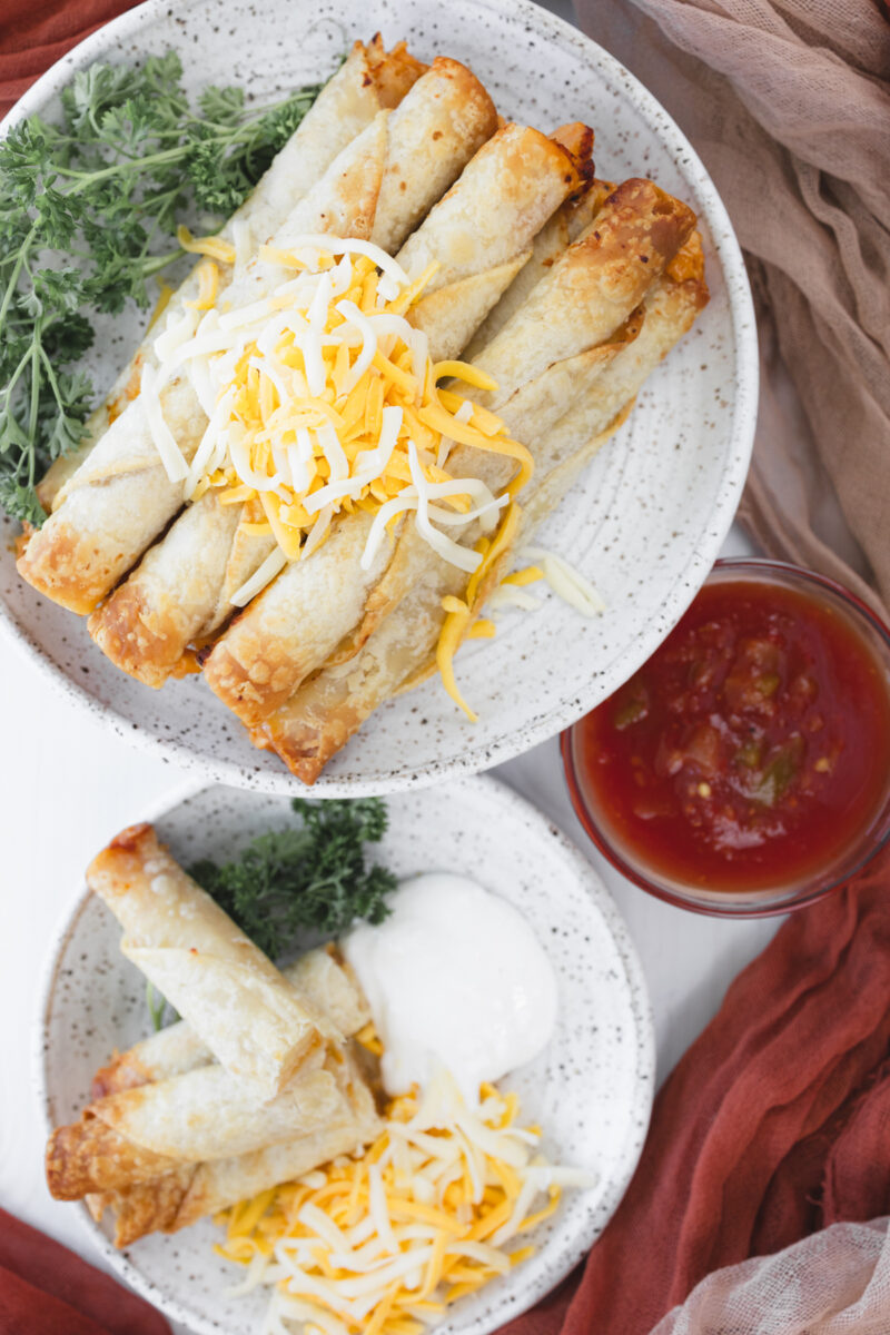 Overhead view of a larger plate stacked with chicken taquitos and shredded cheese, a smaller plate with four taquitos, shredded cheese, sour cream, parsley, and a dish of salsa.