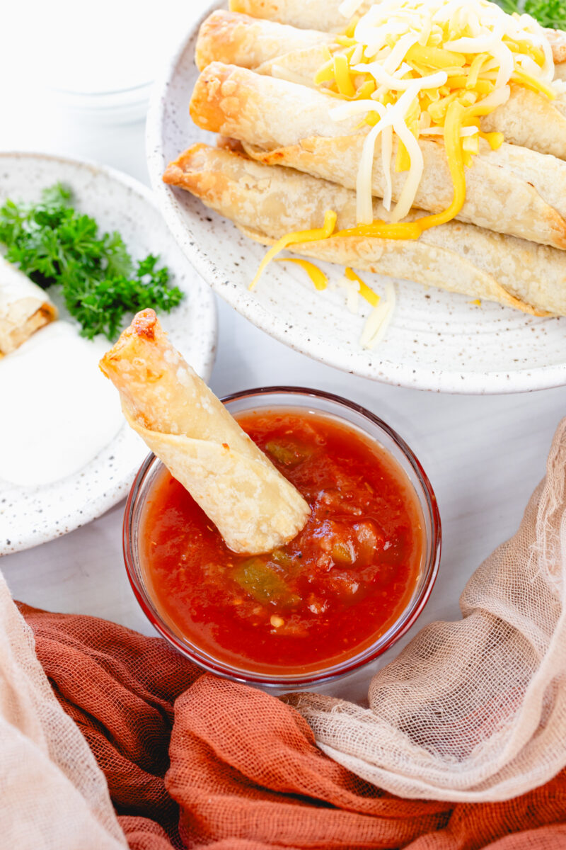 Angled overhead view of a larger plate stacked with chicken taquitos and shredded cheese, a smaller plate with four taquitos, and a salsa dish with a taquito dipped in.