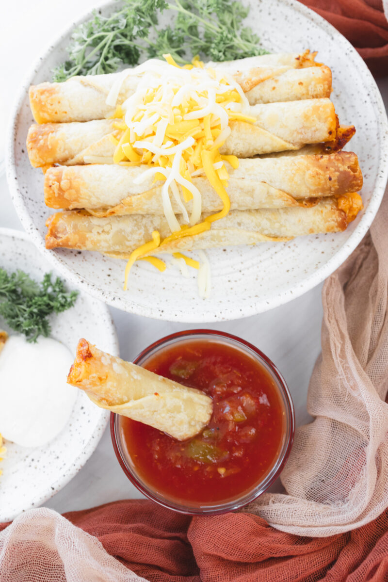 Overhead angled view of a larger plate with stacked chicken taquitos, a smaller plate with four taquitos, and a salsa dish with a taquito dipped in.