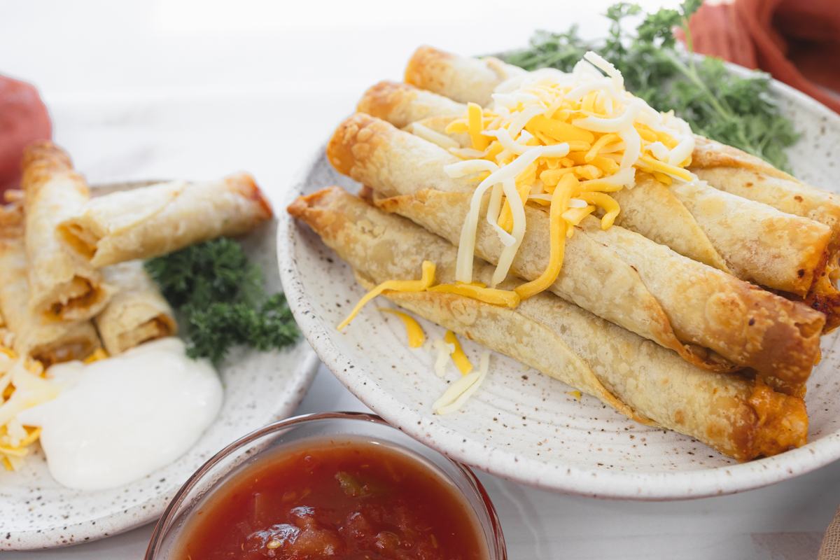 Angled landscape view of a larger plate with stacked chicken taquitos, a smaller plate with four taquitos, and a salsa dish.