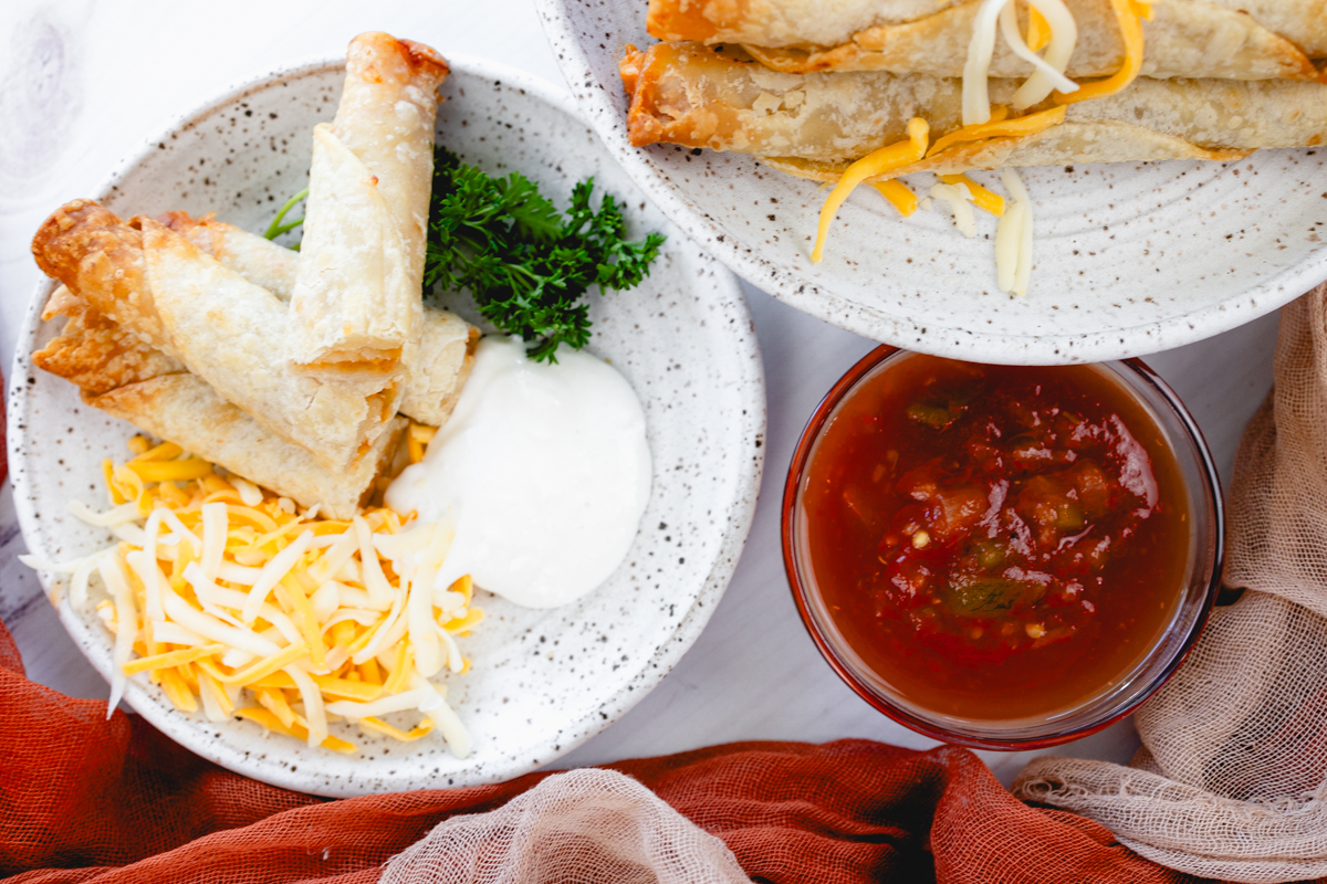 Landscape view of four chicken taquitos on a plate with shredded cheese, sour cream, parsley, next to another plate of taquitos and a small salsa bowl.