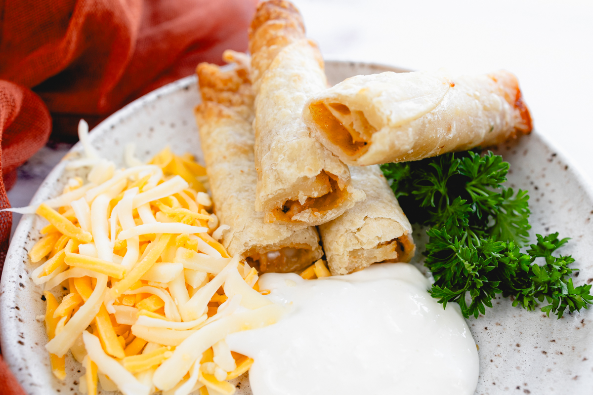 Landscape close-up of four chicken taquitos on a plate with shredded cheese, sour cream, and parsley.