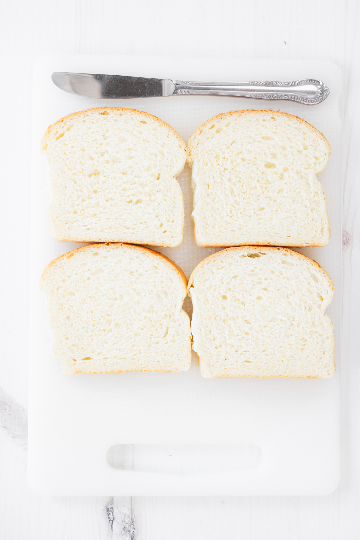Overhead view of four fresh white bread slices placed neatly on a white cutting board next to a butter knife.