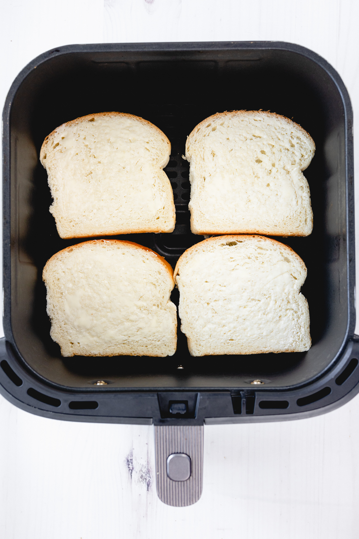 Overhead view of four slices of white bread arranged in an air fryer basket, ready to toast.