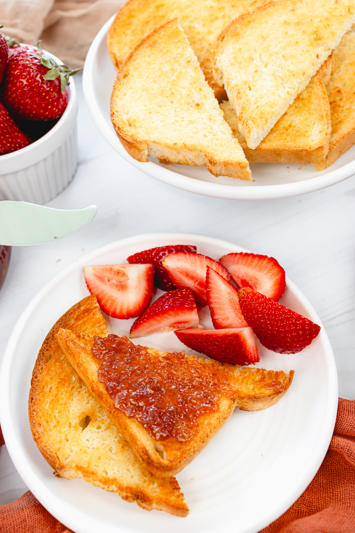 Close-up overhead angled view of golden-brown toasted bread on a white plate with fresh strawberries and jam.