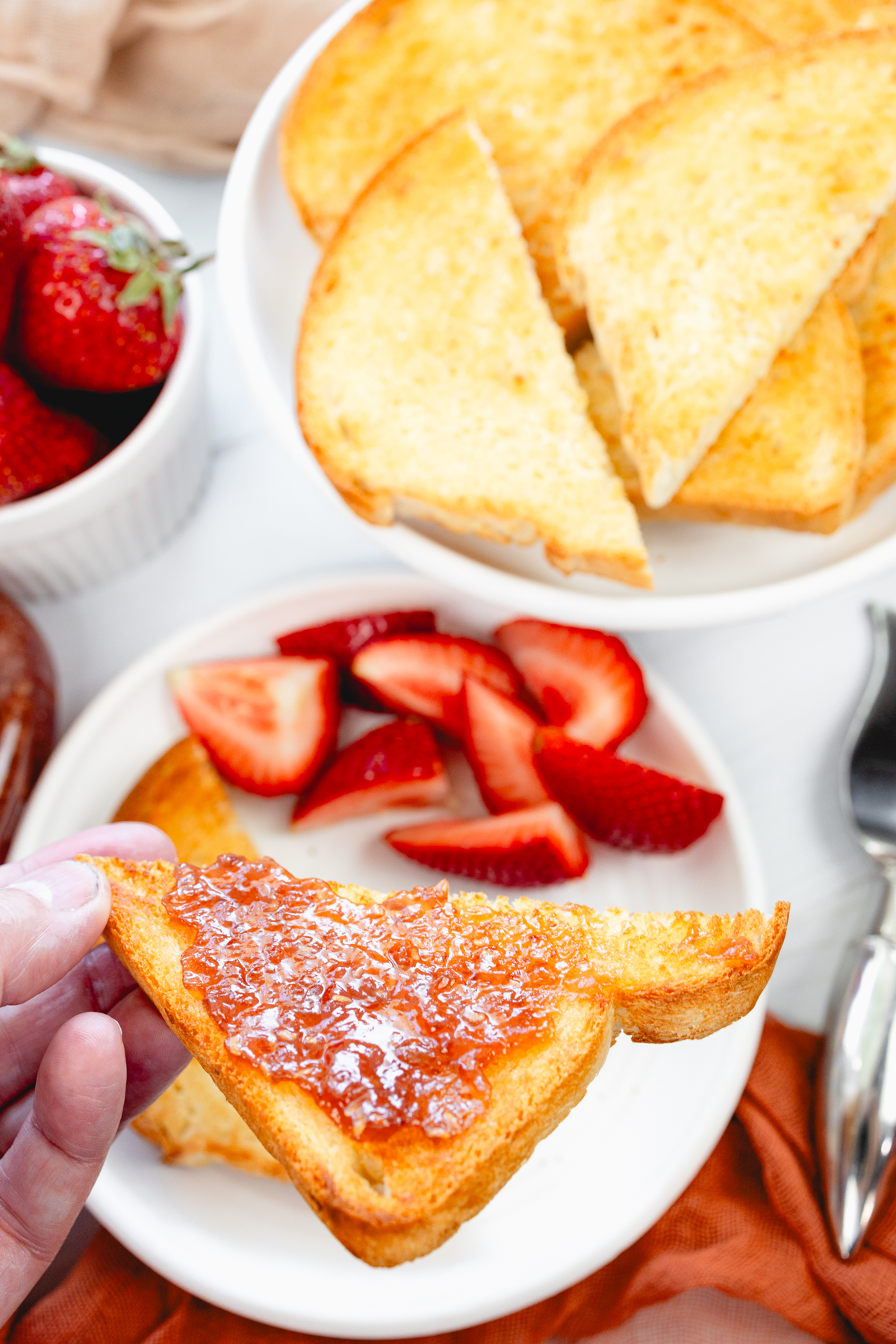 Close-up overhead angled shot of a hand holding a crisp golden slice of toast spread with jam, with strawberries and jam in the background.