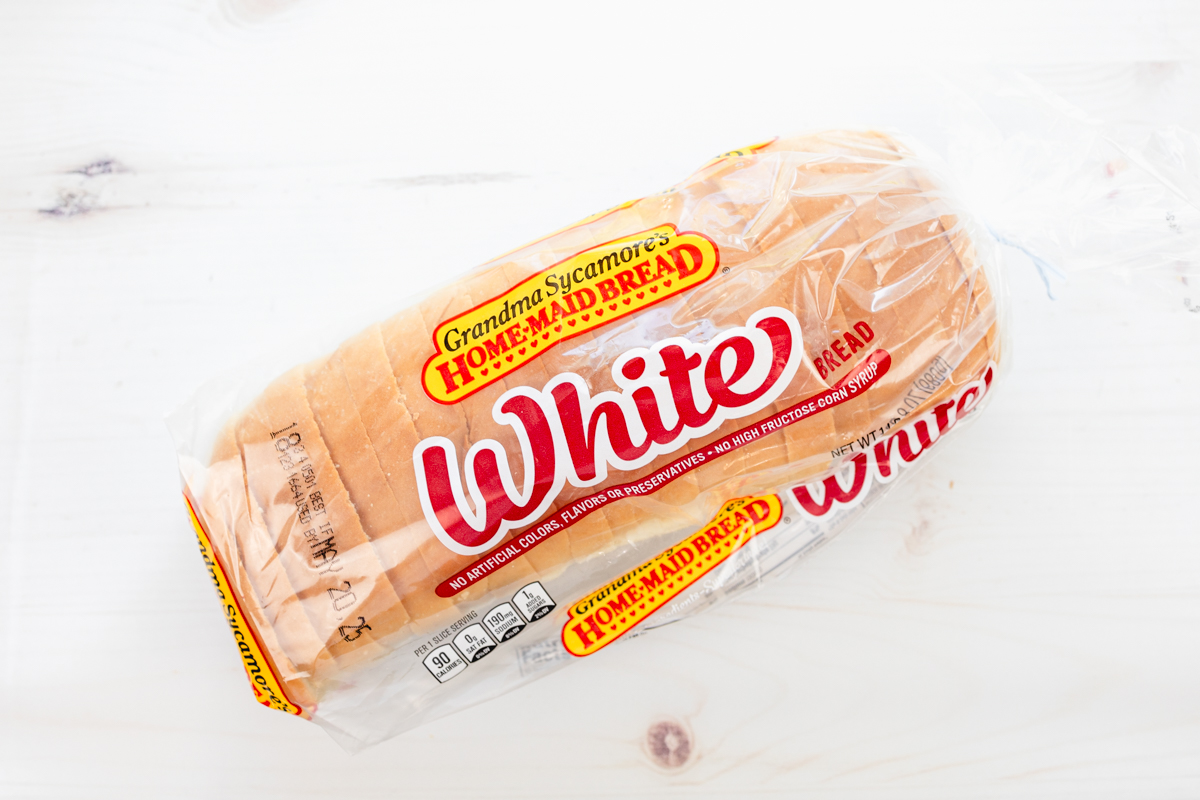 Overhead close-up view of a loaf of sliced white sandwich bread in its original packaging on a white wooden surface.
