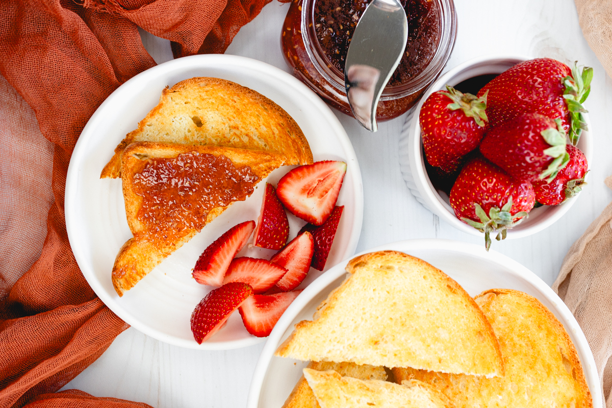 Close-up angled overhead view of golden-brown slices of toast served neatly on a white plate, with strawberries, jam, and a butter knife on the side.