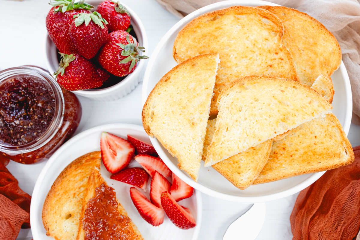 Close-up overhead angled view of golden-brown slices of toast served on a plate alongside fresh strawberries and a small dish of jam.
