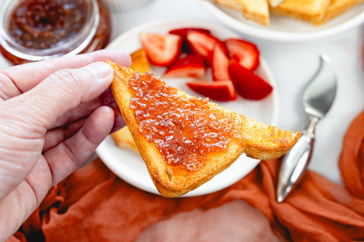 Close-up angled overhead shot of a hand holding one golden, crispy slice of toast to showcase texture, served with strawberries and jam in the background.
